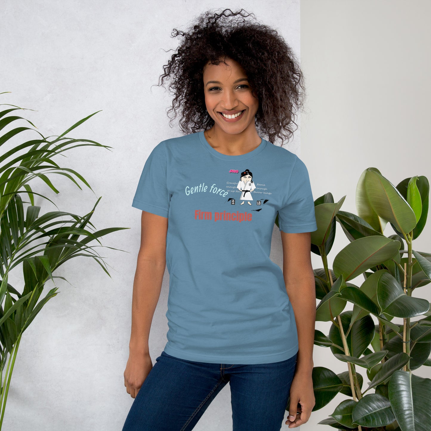 Woman wearing a blue t-shirt with a judo graphic design and text, standing against a white wall with plants.